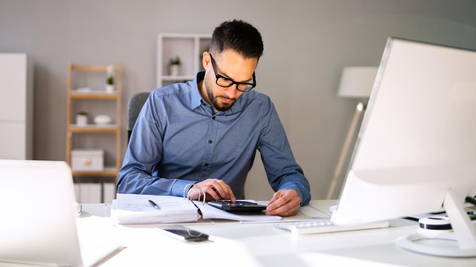 Accountant typing on a calculate at a desk covered with paperwork, a laptop and monitor